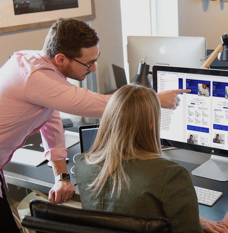 Man pointing at website designs on an iMac screen while a coworker seated at the desk looks on.