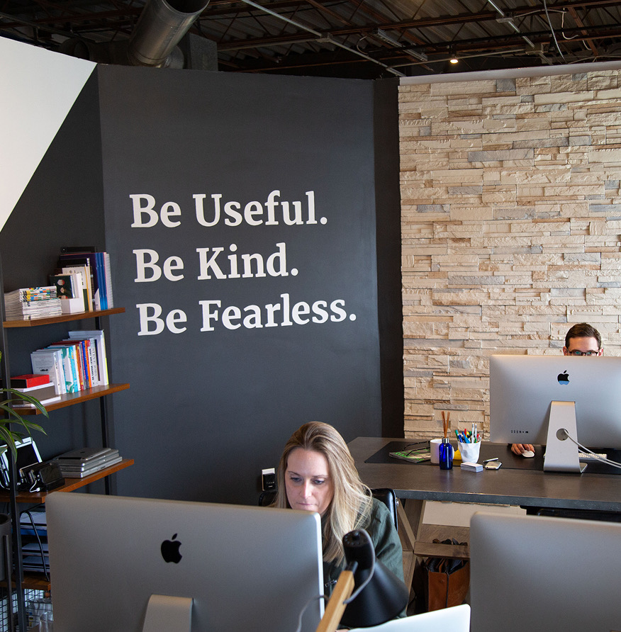 Team members working at iMacs beneath a wall painted with the words Be Useful. Be Kind. Be Fearless.